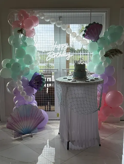 Mermaid-themed balloon arch with pearls, shells and a neon Happy Birthday sign around a cake table